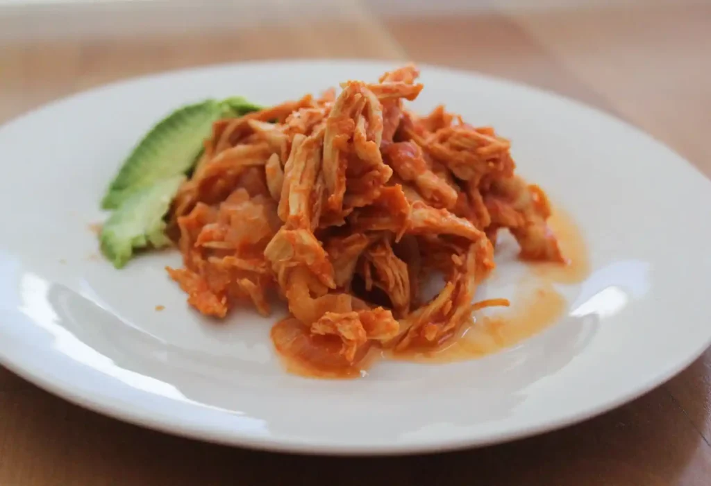 Close-up image of Tinga De Pollo served in a crispy tostada shell, topped with avocado slices and queso fresco